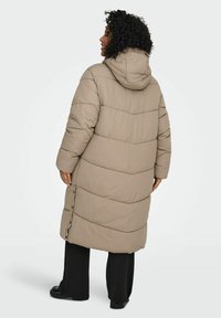 Garš bēšs pūkains mētelis ar kapuci, ar šūtu rakstu, sānu pogām un vaļīgu piegriezumu. Uzvilkts virs melnām biksēm un zābakiem.