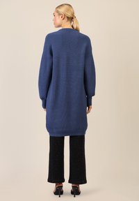 Ylikokoisen sinisen neulejakun ribbitekstuurilla, joissa on pitkät hihat ja pyöreä helma, yhdistettynä mustiin leveisiin housuihin ja korkokenkiin.