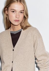 Beige, mjuk cardigan med V-ringning, som har en kn�äppning framtill och ribbade manschetter, lagd över en mörkgrå topp.