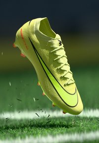 Ghete de fotbal galbene cu un upper texturat, swoosh negru și accente roz. Floate deasupra ierbii verzi, evidențiind un design ascuțit și detalii.