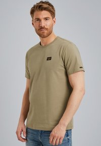 Camiseta de algodón verde oliva con cuello redondo, mangas cortas y un parche de logo negro en el pecho izquierdo. Diseño simple y ajustado.