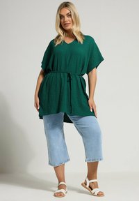 Green textured tunic with a V-neck, cinched waist, and short sleeves, paired with cropped light denim jeans and white sandals.