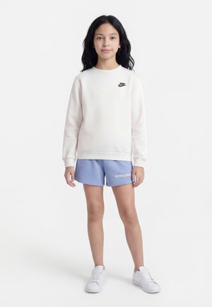 Fille debout portant un sweat-shirt blanc Nike, un short bleu avec le texte "SUNFLOWER" et des baskets blanches sur un fond blanc.