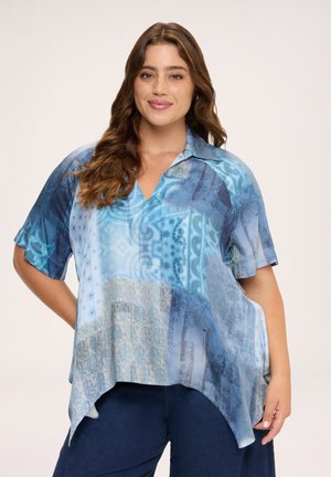 Mujer con cabello largo y castaño que lleva una blusa azul estampada de manga corta y suelta y pantalones azul oscuro, sonriendo con una mano en la cadera.