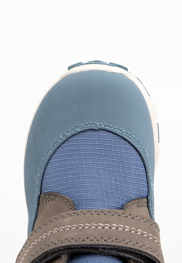 Blauer Textilschuh mit einer glatten, abgerundeten Zehenkappe, ausgestattet mit einem dunkelbraunen Klettverschluss und kontrastierenden Materialien auf der Oberflächenstruktur.
