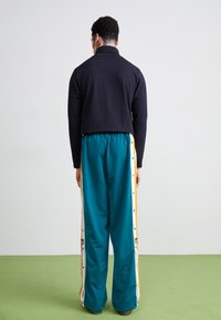 Svart långärmad topp och teal joggingsbyxor med tryckknappar. Byxorna har vita och gula sidostrecker. Slät tygstruktur.