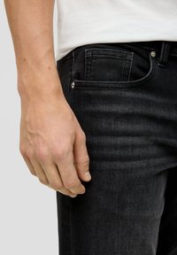 Jean en denim noir avec une coupe slim, comportant une poche avant, un tissu texturé et des garnitures métalliques à la taille. Placement de la main visible près de la poche.