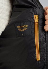 Interior de una chaqueta acolchada negra que presenta un bolsillo con cremallera amarilla y una etiqueta tejida rectangular con "PME LEGEND AMERICAN CLASSIC" en letras doradas.
