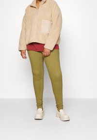 Chaqueta de cremallera beige y suave con un bolsillo crema, superpuesta a una camisa burdeos. Pantalones ajustados verde oliva y zapatillas de plataforma blancas completan el atuendo.