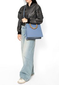 Borsa in pelle blu con manico in bambù, forma rettangolare e angoli netti, abbinata a una giacca bomber nera e jeans a gamba larga.