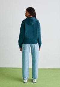 Hoodie turquoise avec une poche kangourou avant et un cordon de serrage, associé à un jean large bleu clair. Baskets blanches avec accents verts.