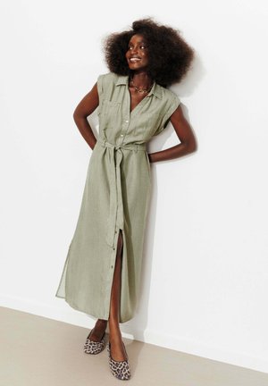 Femme aux grands cheveux bouclés portant une robe verte sans manches boutonnée avec une ceinture à la taille et des ballerines imprimé léopard, appuyée contre un mur blanc.