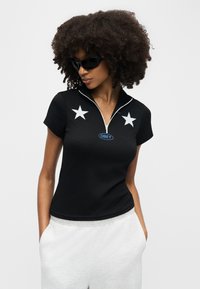 Γυναίκα με μεγάλα σγουρά μαλλιά και μαύρα γυαλιά ηλίου, φοράει μαύρο πουκάμισο με φερμουάρ με λευκά αστέρια και λογότυπο "OBEY", τα χέρια στις τσέπες λευκών παντελονιών.