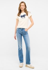Camiseta crema con un gráfico de caballo azul marino y letras, combinada con jeans de color azul claro. Zapatillas texturizadas con acentos beige completan el look.