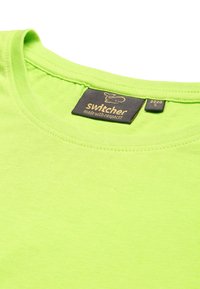 T-shirt in cotone verde neon con scollatura rotonda e texture morbida. L'etichetta visibile mostra il marchio "Switchers" e la taglia S.