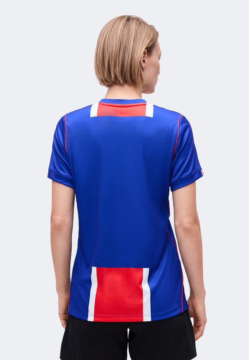 Personne aux cheveux blonds courts portant un maillot de sport bleu avec des rayures verticales rouges et blanches dans le dos, associée à un short noir, vue de dos.