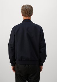 Man with short hair wearing a plain dark navy bomber jacket and black pants, standing facing away against a plain light background.