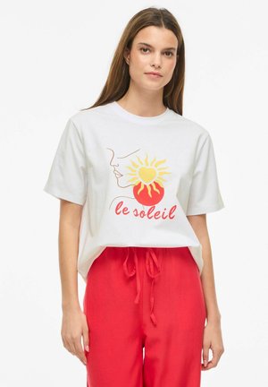 Femme portant un t-shirt blanc avec un dessin en lignes du soleil, du cœur et du visage et un texte rouge "le soleil", associé à un pantalon rouge à cordon.