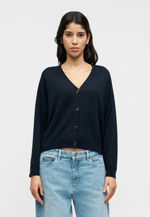 Πλεκτή ζακέτα σε ναυτικό μπλε χρώμα, κατασκευασμένη από απαλό ύφασμα, με V-λαιμόκοψη, μακριά μανίκια και πέντε σκουρόχρωμα κουμπιά στο μπροστινό μέρος. Κοντό σχέδιο.