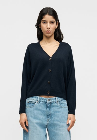 Cardigan bleu marine en tissu tricoté doux, avec un col en V, des manches longues et cinq boutons foncés sur le devant. Design court.