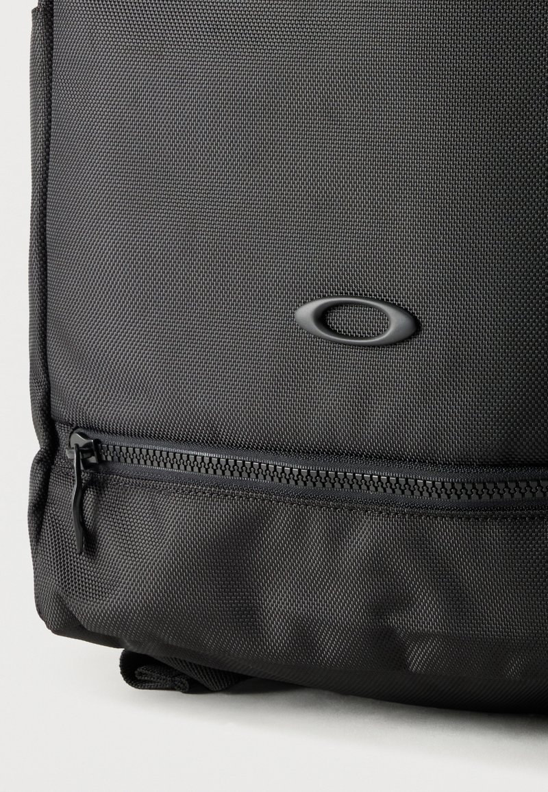 Gros plan sur un sac en tissu noir texturé avec une fermeture éclair horizontale et un logo ovale centré au-dessus de la fermeture éclair.