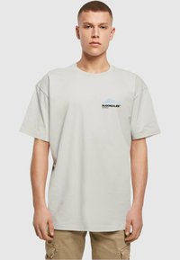 Lys grå oversized t-shirt lavet af bomuld, med et lille sort og blåt logo på venstre bryst. Korte ærmer og rund halsudskæring.