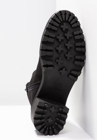 Semelle de botte noire avec un motif de crampons profonds comprenant des crampons en forme de croix et rectangulaires, fermeture éclair latérale visible sur la partie supérieure.