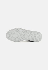 Semelle d'une basket blanche avec des motifs géométriques texturés et un logo au centre, présentée sur un fond clair uni.