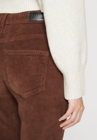 Bruine corduroy broeken met een verticale ribbelstructuur, voorzien van achterzakken en een zwart leren merklabel bij de taille. Witte truiarm zichtbaar.