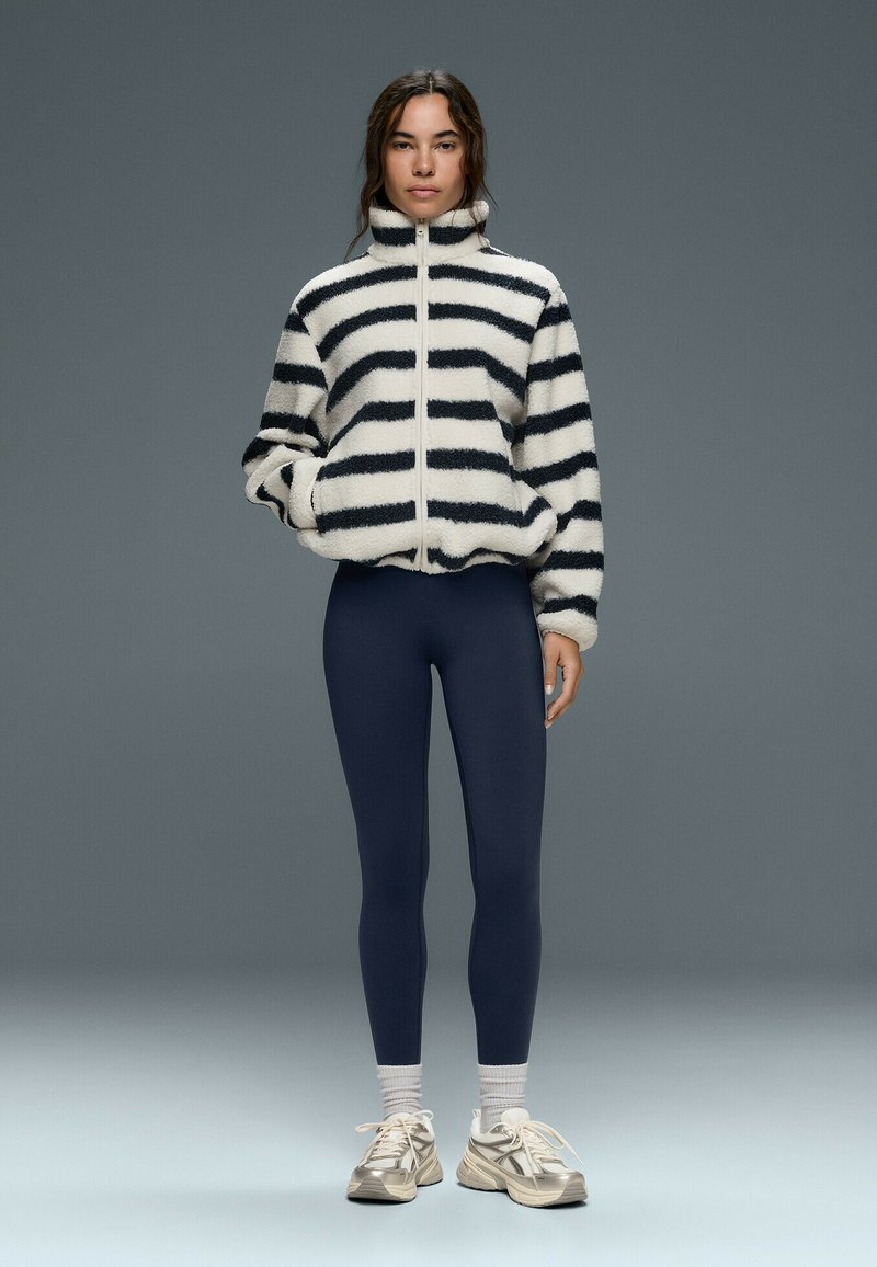 Femme debout portant une veste polaire zippée rayée noire et blanche, des leggings bleu marine, des chaussettes blanches et des baskets beige sur fond gris.