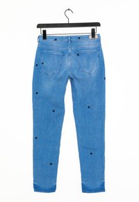 Vaqueros de mezclilla azul con patrones de estrellas de color azul marino bordados, corte ajustado, cinco bolsillos y una etiqueta en la pretina. Se cuelga en una percha negra sobre un fondo blanco.