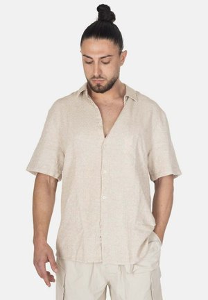Uomo con capelli scuri raccolti in uno chignon indossa una camicia beige chiaro a maniche corte e pantaloni chiari, guarda in basso con una mano in tasca.