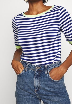 Femme portant une chemise rayée bleu et blanc avec des bordures vertes et un jean bleu taille haute, les mains dans les poches.