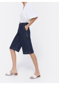 Mujer con blusa blanca de mangas abullonadas, pantalones cortos de mezclilla oscuros hasta la rodilla con bolsillos laterales y sandalias de tacón bajo de punta abierta blancas, caminando hacia la derecha.