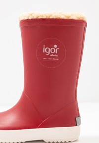 Botte de pluie en caoutchouc rouge avec une doublure en polaire crème sur le dessus, semelle texturée blanche et un accent gris au niveau du talon. Étiquette de marque visible.