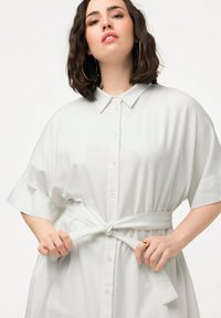 Biała, oversize koszula na guziki z kołnierzykiem i paskiem w talii, z krótkimi rękawami i gładką teksturą materiału.