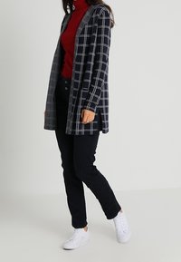 Rutig cardigan i marinblått och grått, lager på lager över en röd polotröja. Kombineras med mörka byxor och vita sneakers, med sidoklipp och knapppdetaljer.