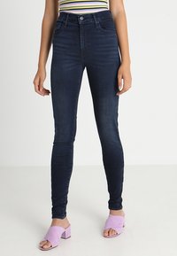 Tummansiniset kapeat farkut korkealla vyötäröllä, joissa on sileä denim-kangas, etutaskut ja kontrastisaumaukset.