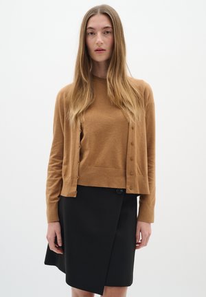 Hnedý cardigan s gombíkmi vyrobený z jemného materiálu, skombinovaný s čiernou zaviazanou sukňou s bočným gombíkovým detailom; hladká textura, jednoduchý dizajn.