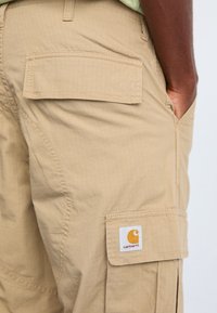 Beige Cargo-Shorts aus leichtem Stoff, mit zwei großen Seitentaschen, einem strukturierten Rautenmuster und einem weißen Carhartt-Label mit orangefarbenem Logo.