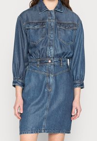 Denimmekko, jossa on pitkät puff-hihat, nappikaulus, kaksi rinta-taskua ja vyötärön kiristyminen vyöllä keskipesussa.