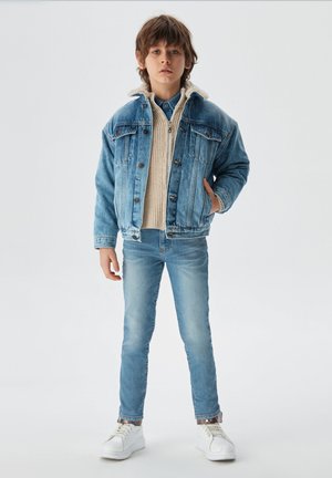 Jeansjacke mit einer hellen Waschung, Kragen aus Fleece, kombiniert mit einem beigen Strickpullover und hellblauen Slim-Fit-Jeans. Weiße Sneaker.
