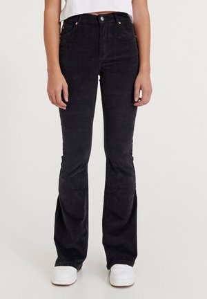 Personne portant un pantalon flare en velours côtelé noir taille haute avec des poches avant et des baskets blanches, se tenant devant un fond uni.