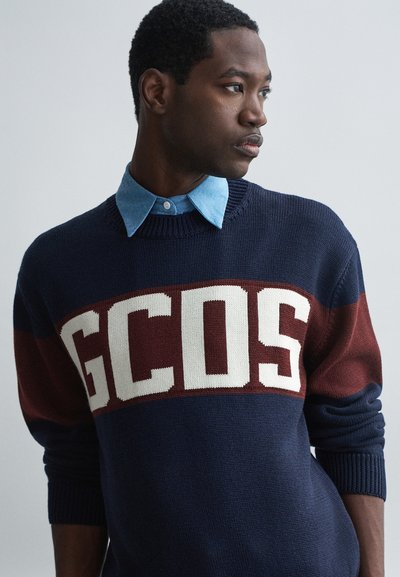 Tumesinine kudumipusa, millel on tumepruun horisontaalne triip ja valge "GCOS" logo. Kandmine helesinise kraega särgi peal. Tekstuuriga kangas.