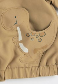 Veste beige avec une appliqué de dinosaure couleur fauve, un ventre blanc, des contours bleus et des taches, fabriquée à partir d'un tissu lisse au fini texturé.