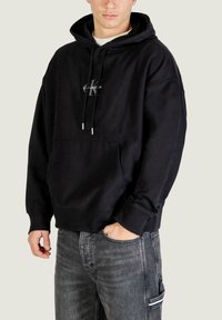 Sudadera negra con capucha y bolsillo frontal, con un pequeño logo en el pecho, hecha de tela suave, diseño holgado y detalles con cordones.