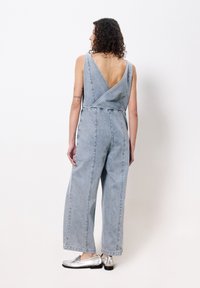 Denim overall in lichtblauw met een V-rug en brede pijpen. Met een wikkelstijl buste en zichtbare stiksels. Draagt met zilveren schoenen.