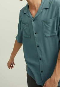 Uomo che indossa una camicia a maniche corte color turchese con due tasche sul petto, che estende il braccio sinistro verso il basso, su uno sfondo chiaro e semplice.