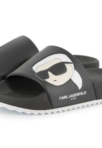 Sandales noires à glisser avec une tige lisse présentant un personnage de dessin animé en blanc, portant des lunettes de soleil. Le texte sur la semelle indique "KARL LAGERFELD KIDS."