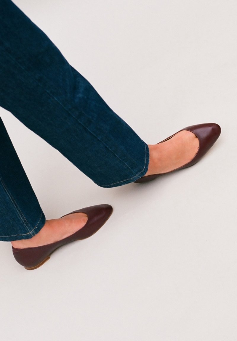 Pieds portant des ballerines à bout pointu de couleur brun foncé associées à un jean denim bleu foncé sur un fond clair.